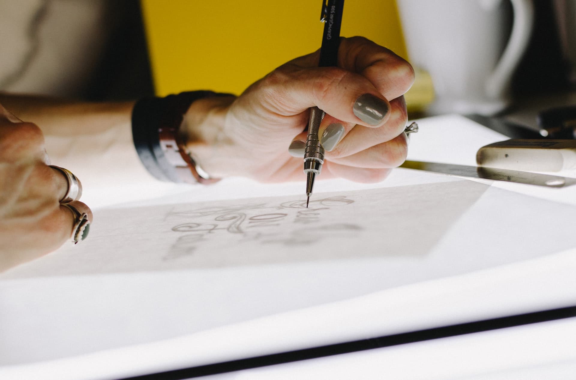 A hand with painted nails grasps a digital stylus mid-stroke atop a bright white screen with a creative document displayed