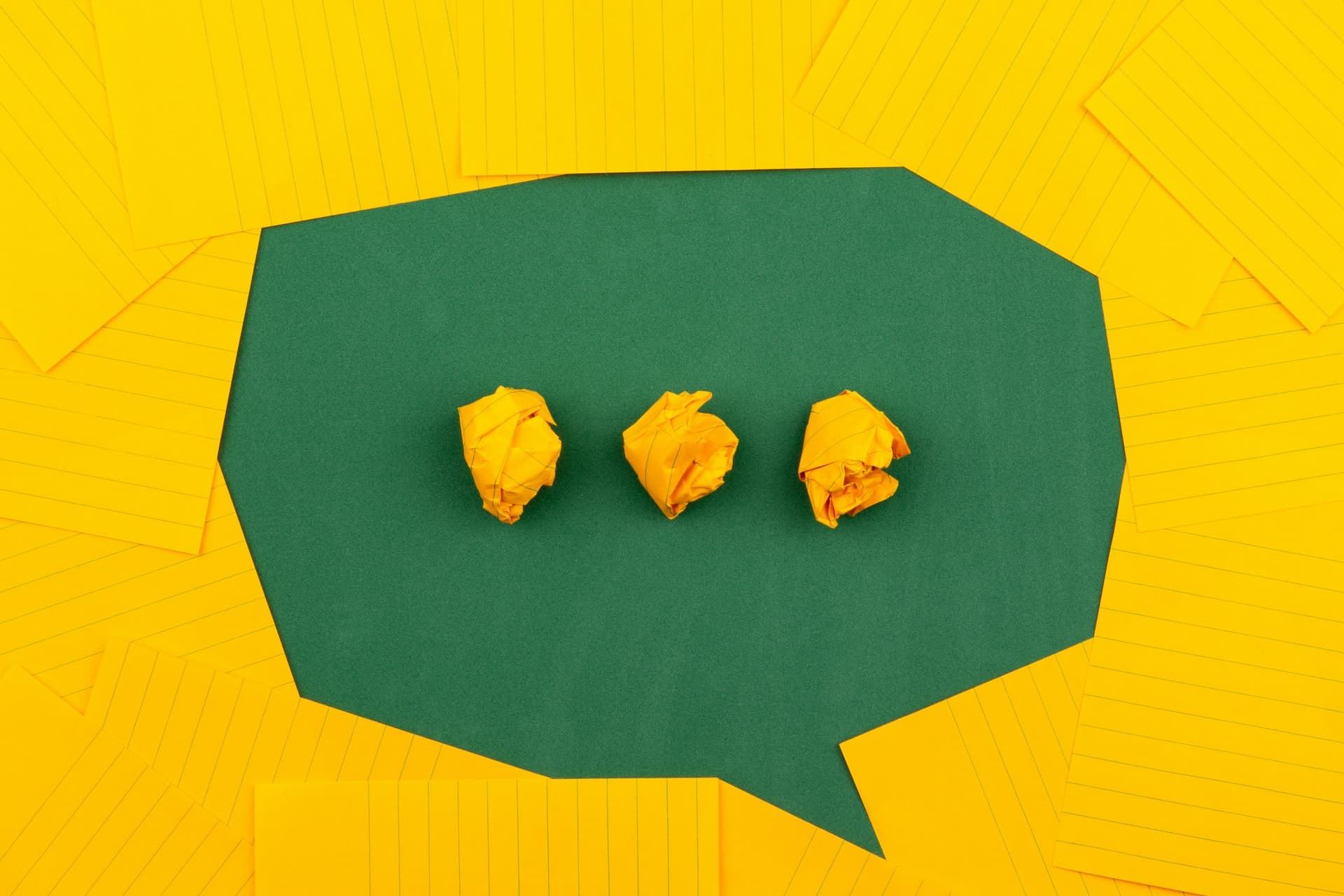 A speech bubble and out of yellow lined paper arranged on a green wall. Three crumpled up balls of yellow paper form three dots inside the speech bubble.