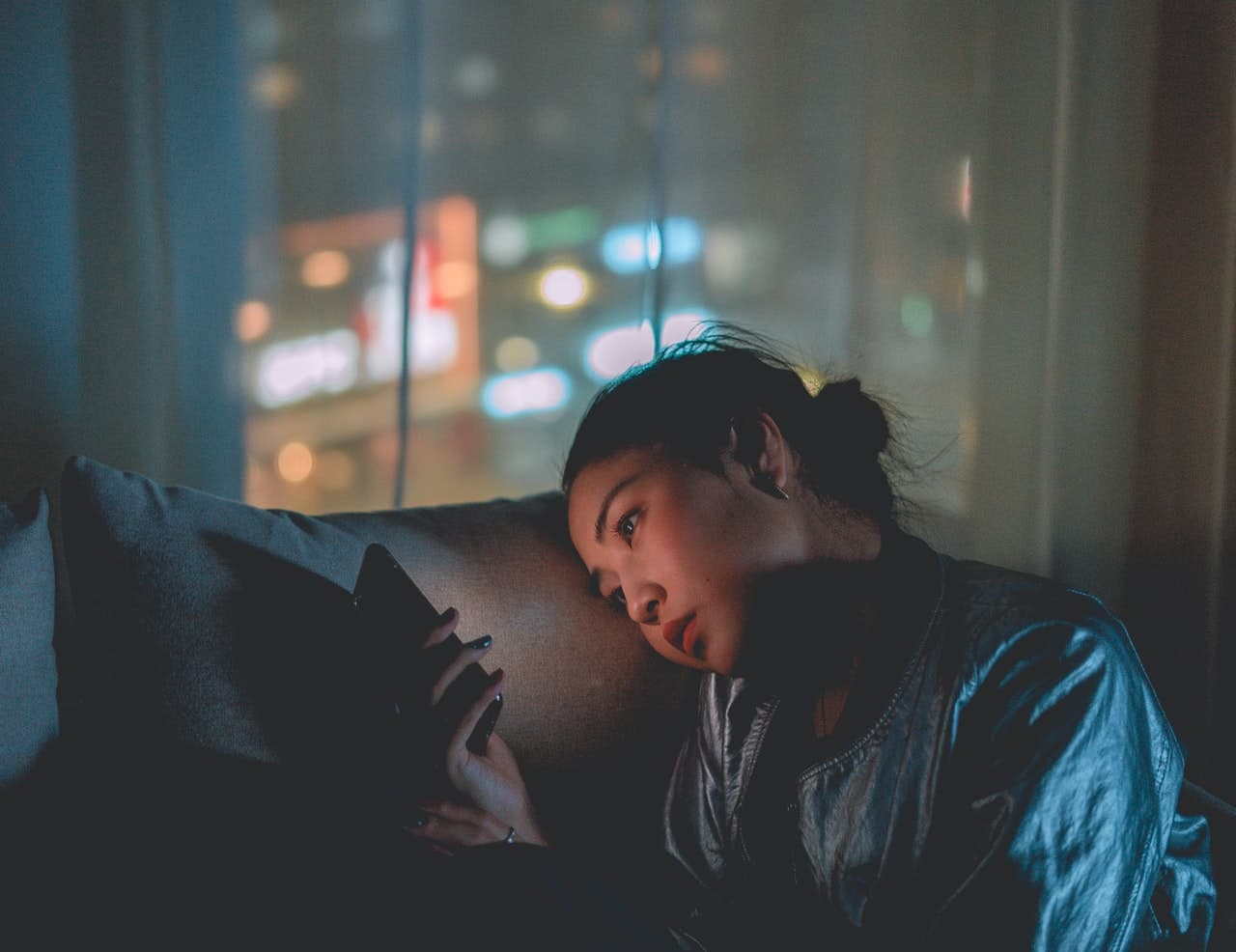 A femme person laying on the couch with their face rested on a cushion and illuminated by the glow of their phone in a dark room. Some neon lighting from street outside is visible through a curtain.