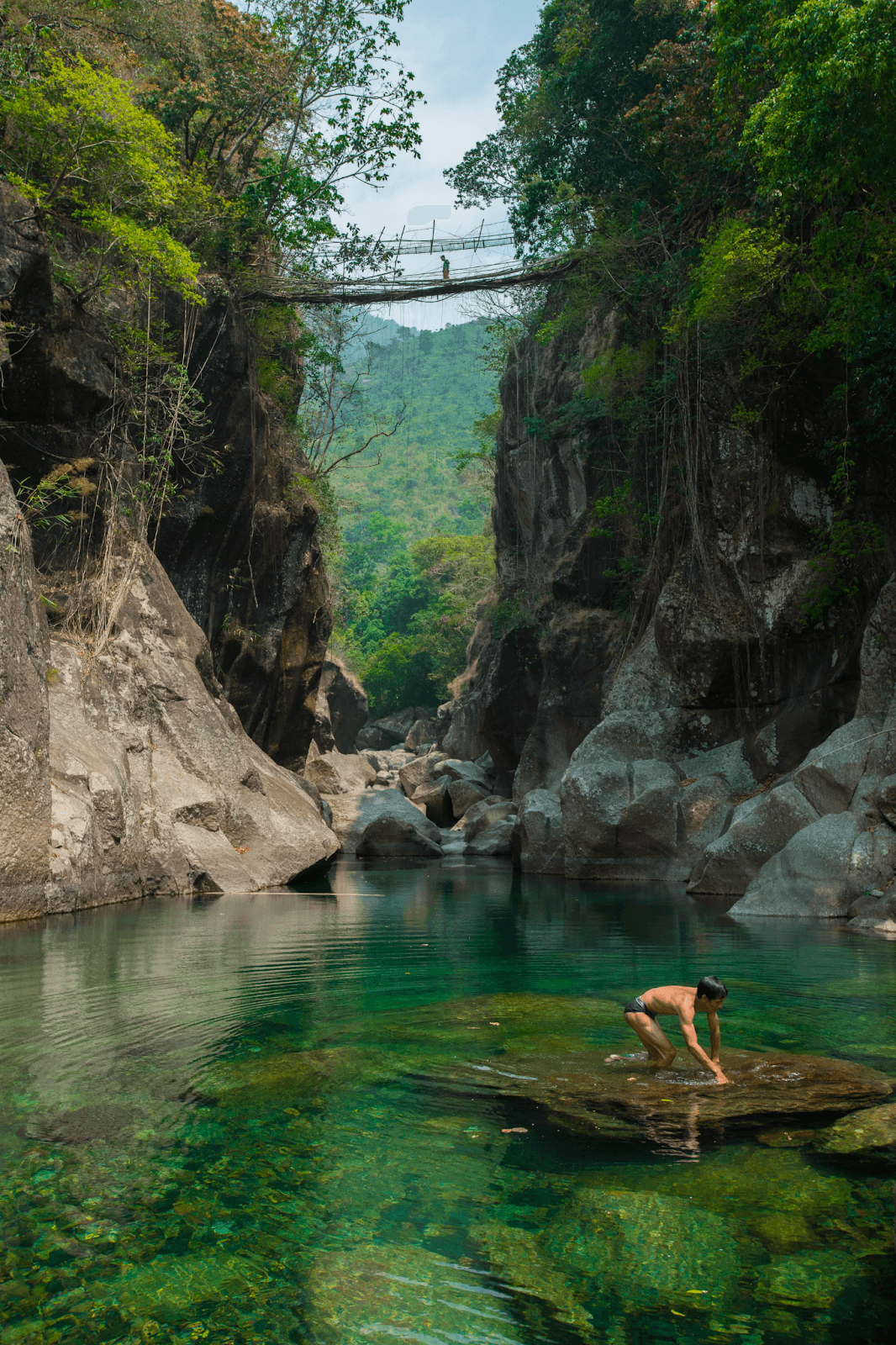 A person is crossing a bridge made of roots that connects two cliffs above a river. Down in the river, a man takes a bath.