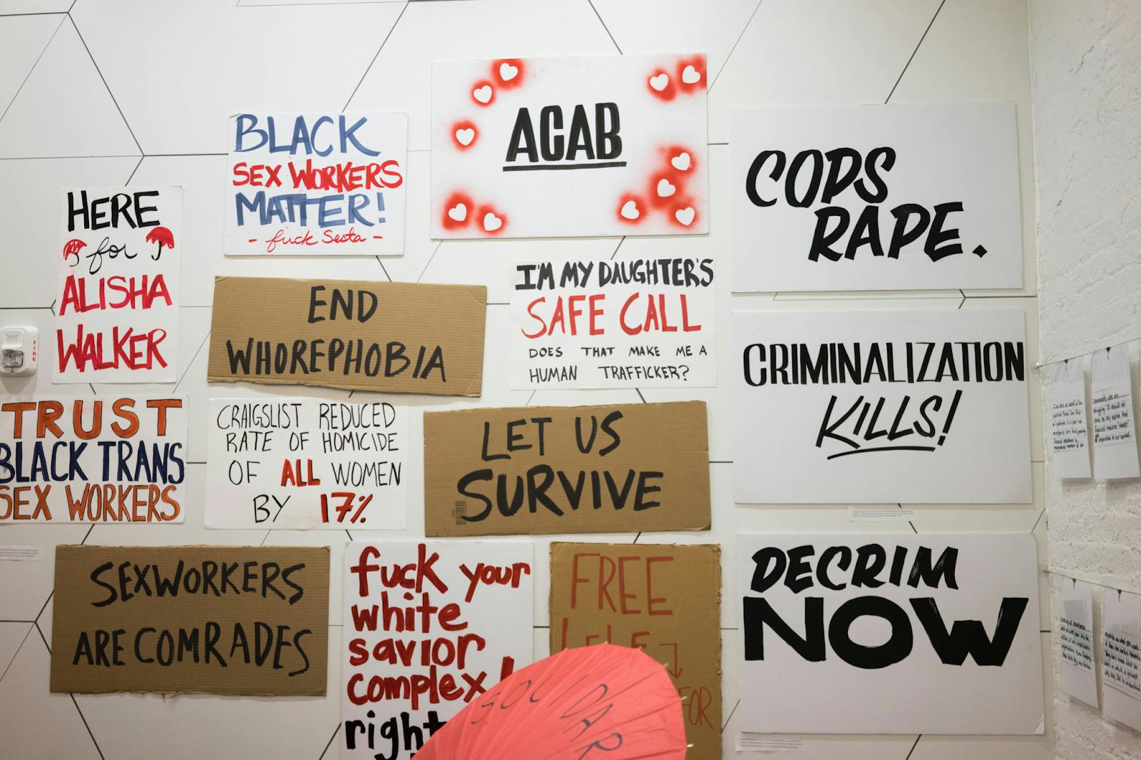 Fourteen protest signs on poster board and cardboard, featuring red and black lettering, are mounted and arranged on a white wall. Their messages range from calls to free incarcerated sex workers and to protect and trust Black trans sex workers, to declaratives against the police and criminalization.