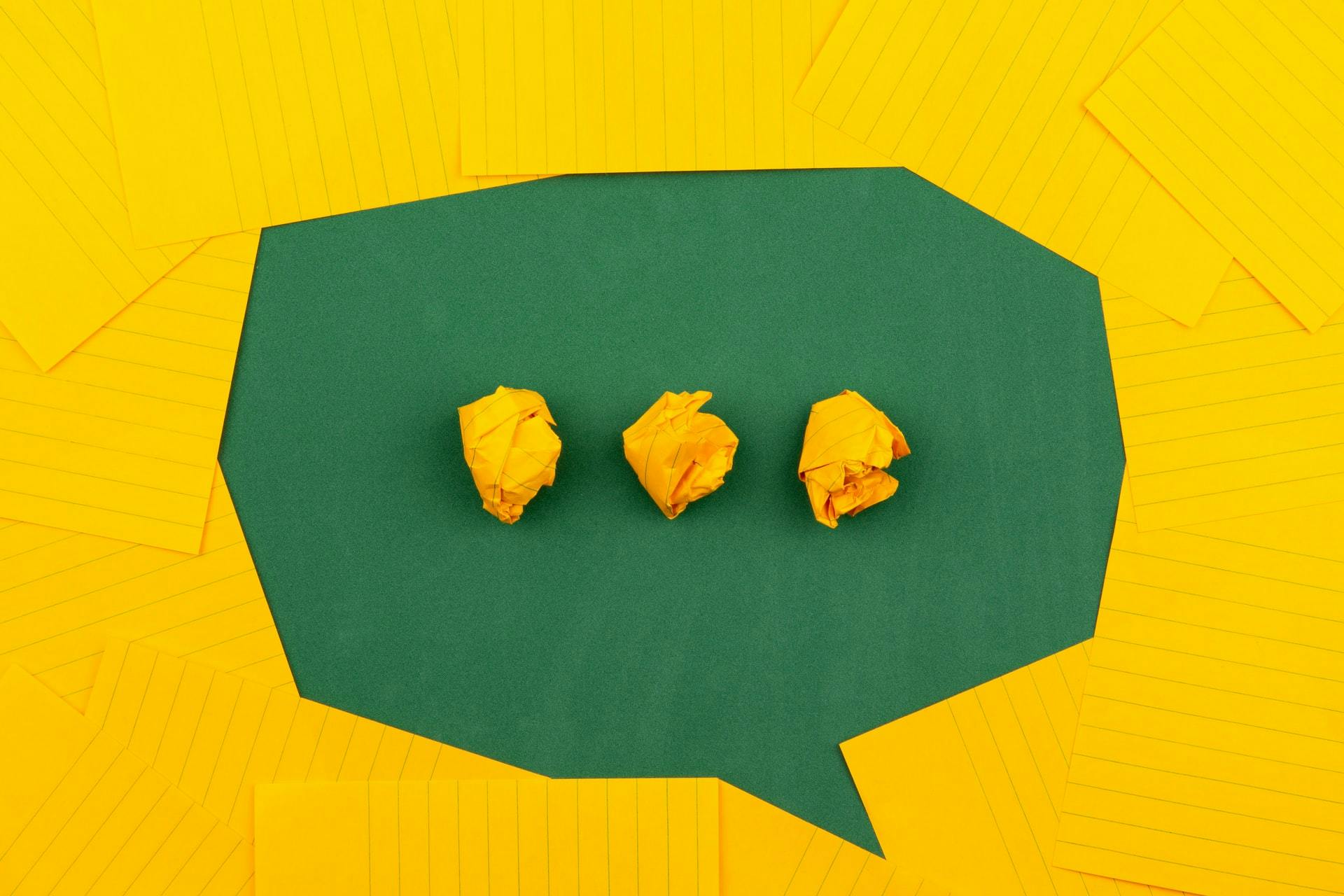 A speech bubble and out of yellow lined paper arranged on a green wall. Three crumpled up balls of yellow paper form three dots inside the speech bubble.