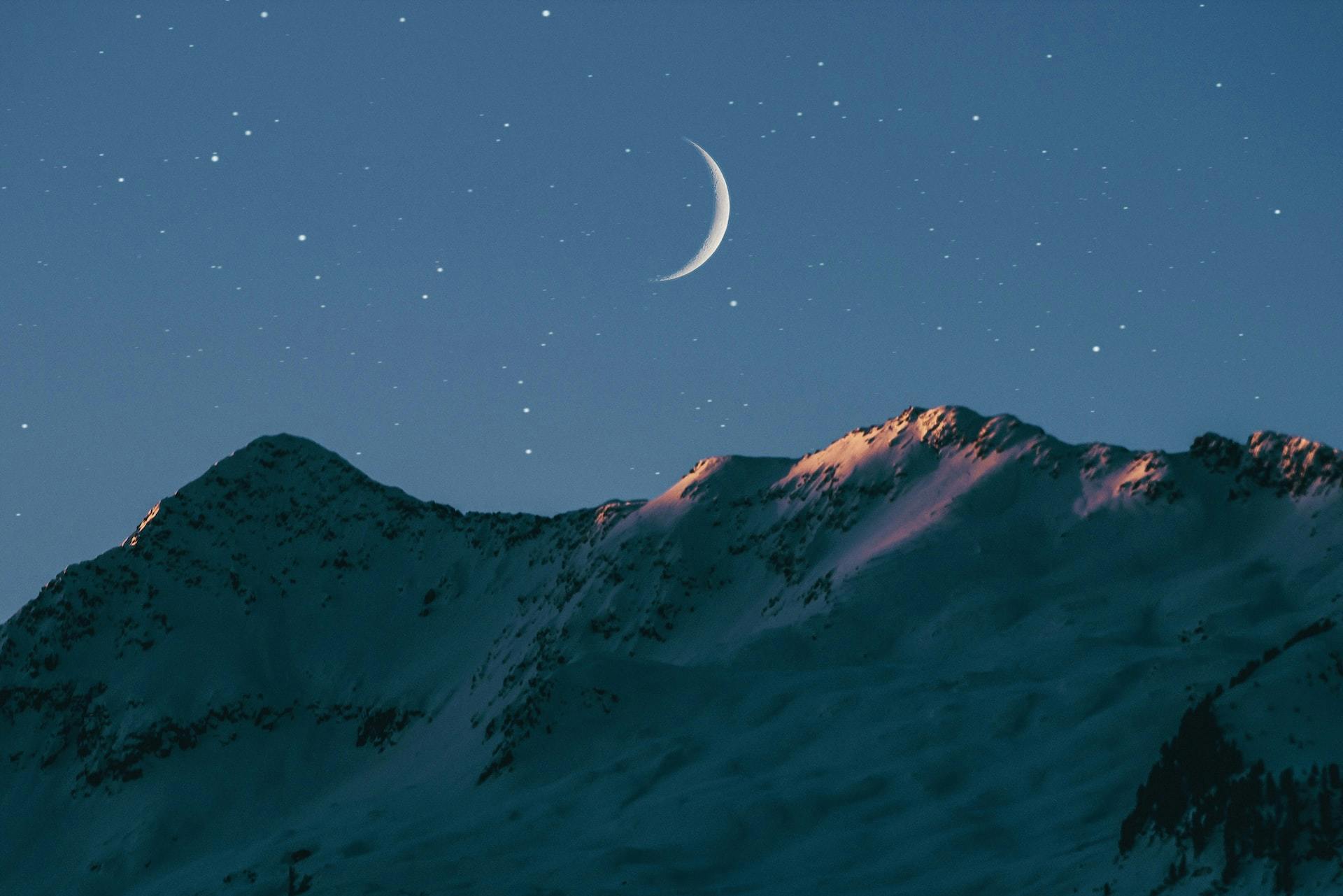 The night sky with stars and a crescent moon visible above snowy mountains whose peaks are just illuminated by the setting sun
