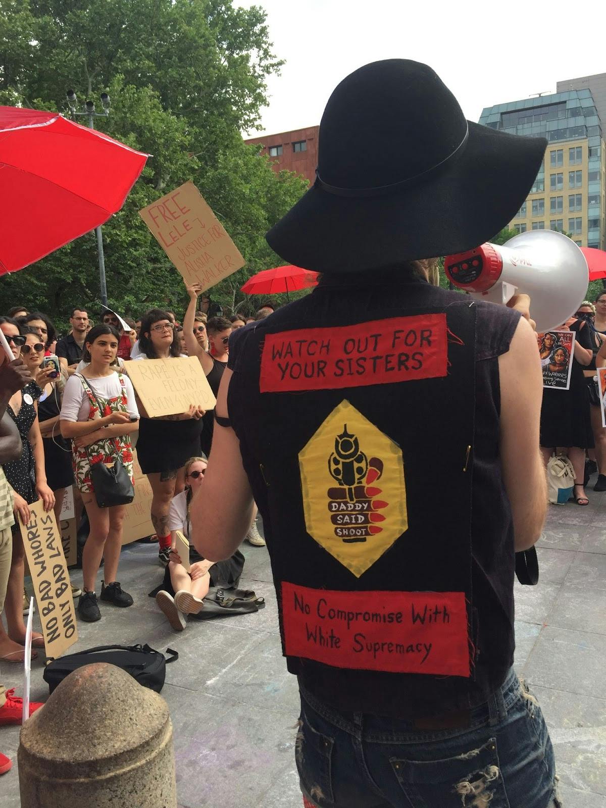 A person stands with their back to a crowd at a protest. They are holding a megaphone and wearing a black vest. The vest says, "Watch out for your sisters. No compromise with white supremacy."