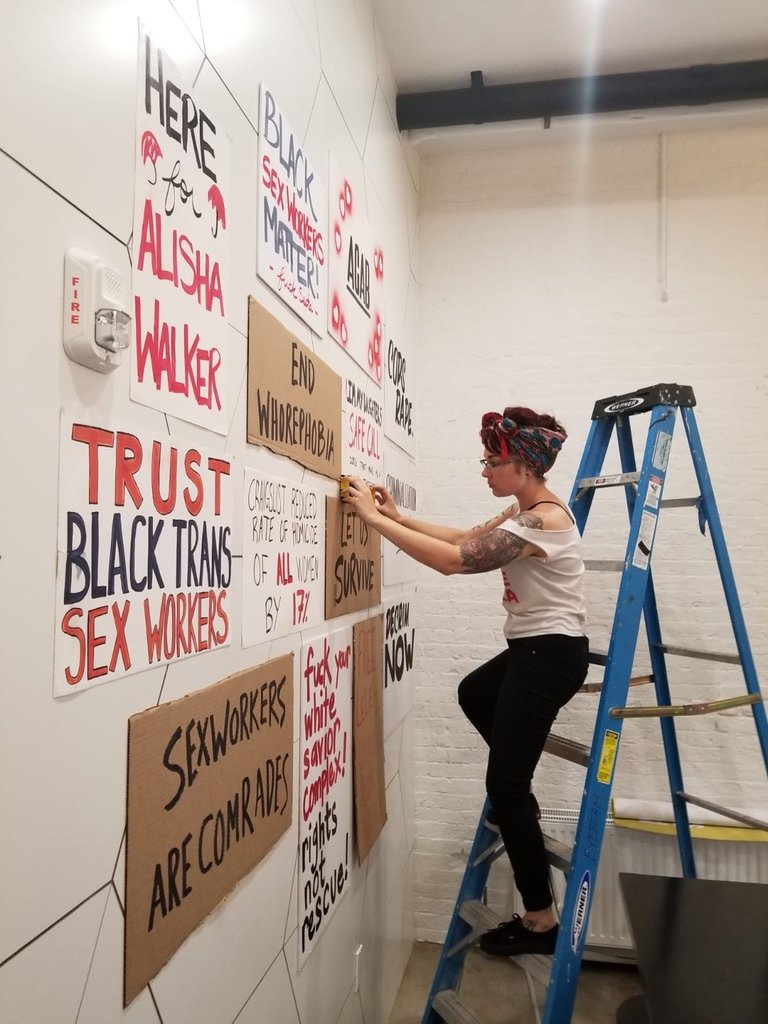 A white, tattooed femme is perched on the steps of a ladder adjusting protest signs they’ve mounted to the white wall across from them. The signs feature black and red lettering spelling out messages of love, solidarity, and rage from sex working community.