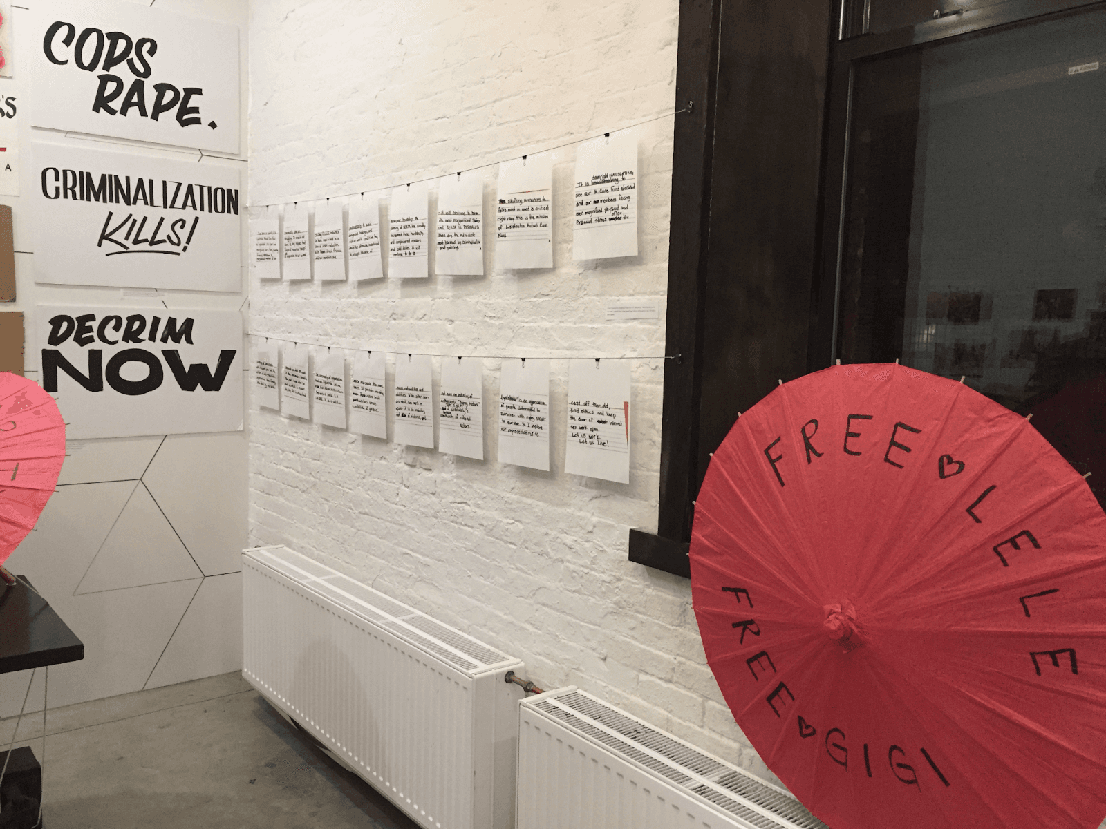 An opened red parasol featuring the words “Free LeLe” and “Free GiGi” written in black marker is mounted on a windowsill. To the parasol’s left are two rows of metal twine, with enlarged printouts of a speaker’s notecards, attached with black alligator clips. Part of another red parasol atop a table and posters from the last two reflections are visible further left in the image.