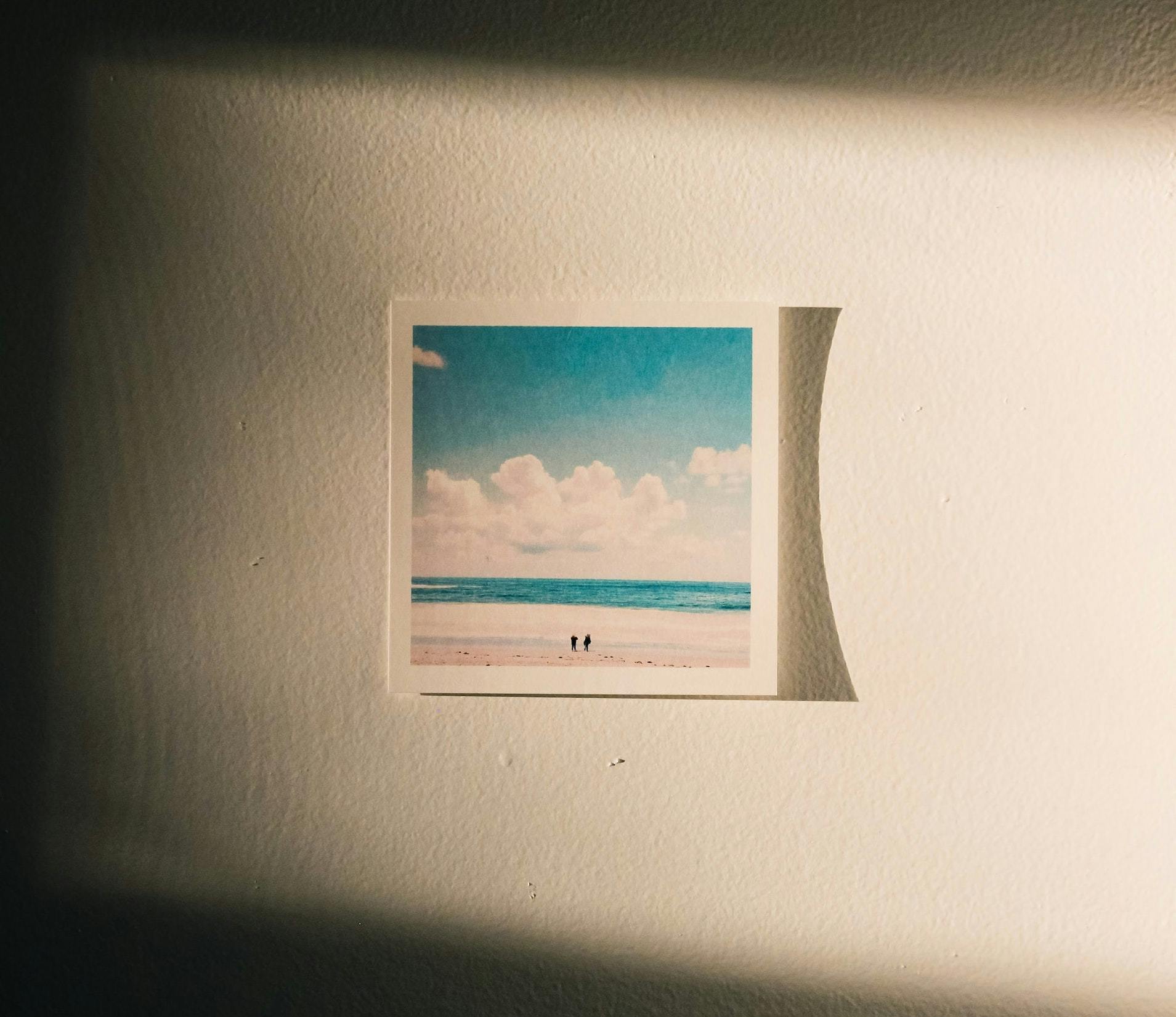 A square printed photograph or artwork of a beach where two small figures are visible is illuminated by warm light streaming through a window onto a wall